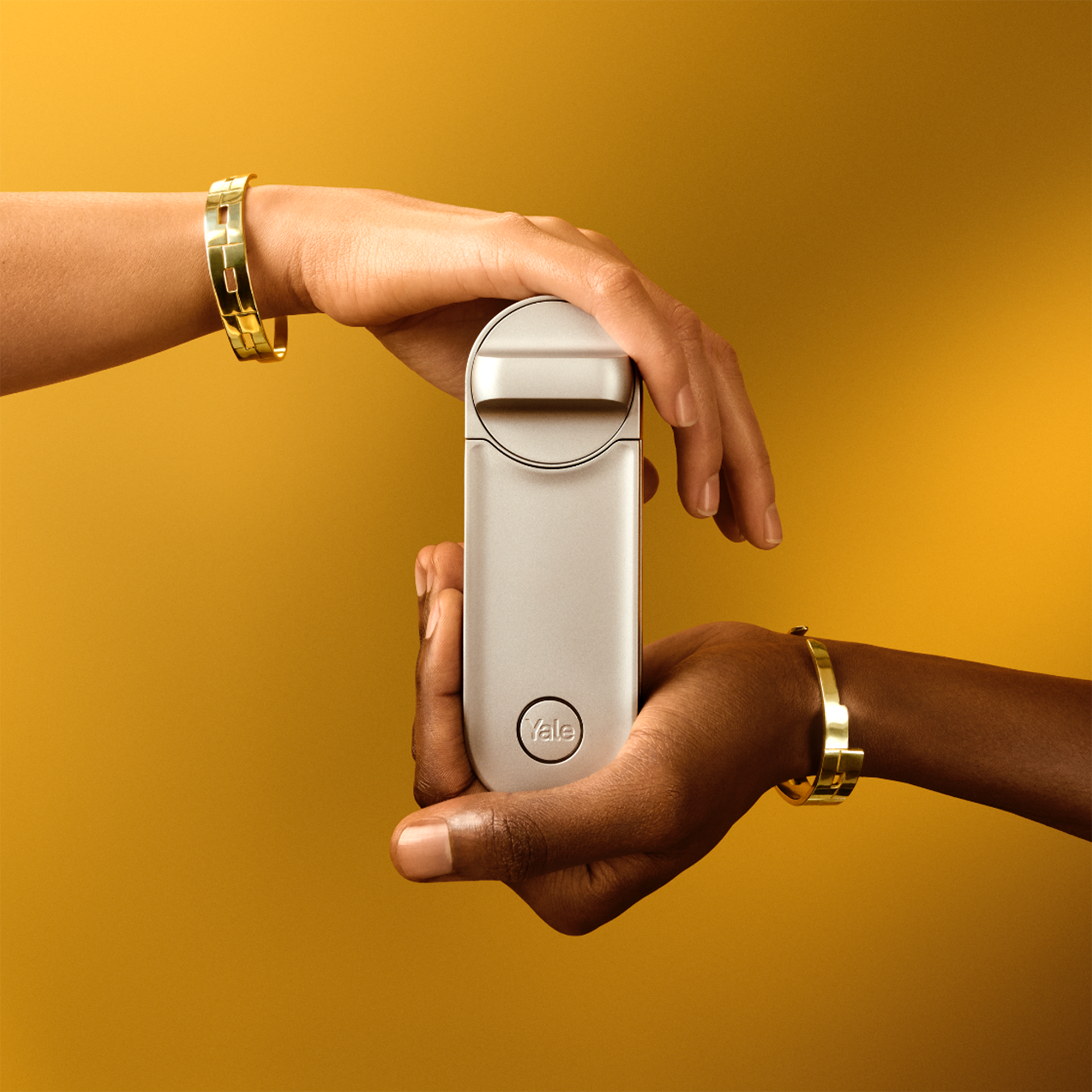 Zwei Hände mit goldenen Armbändern halten ein silbernes Linus L2 Smart Lock, eine Hand oben und eine unten, vor einem goldenen Hintergrund.